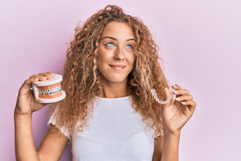Smiling woman with curly hair and brown eyes holding clear aligners in one hand and a braces model in the other, comparing orthodontic options.