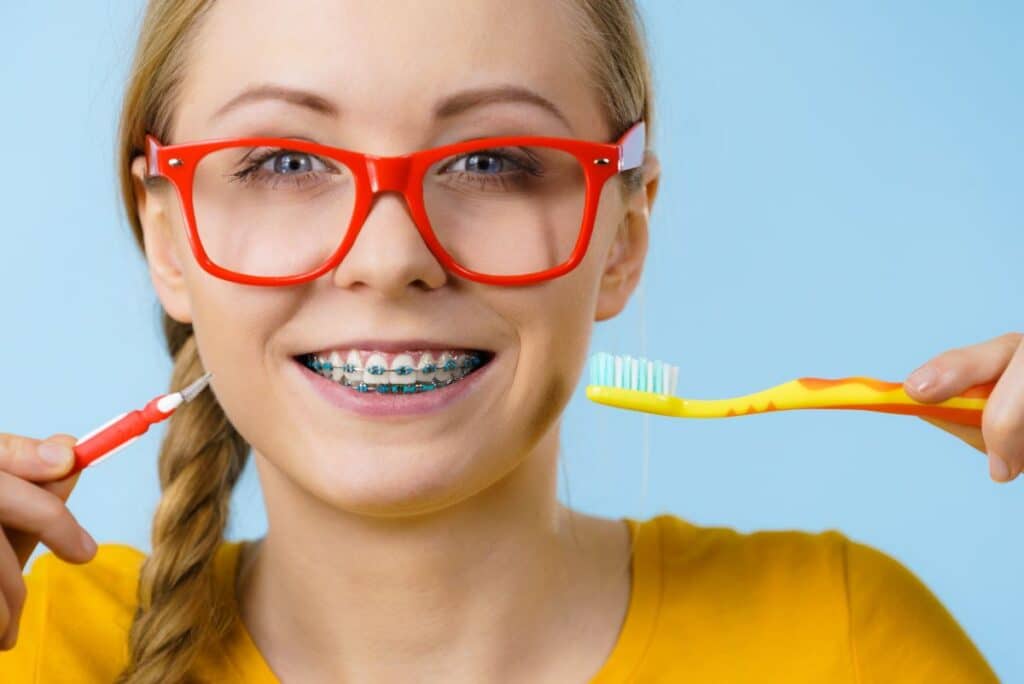 A young person wearing braces and red glasses holds a toothbrush in one hand and a small interdental brush in the other.