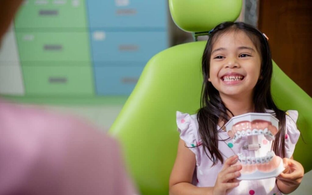 Young girl smiling while holding a dental model with braces during an orthodontic consultation.