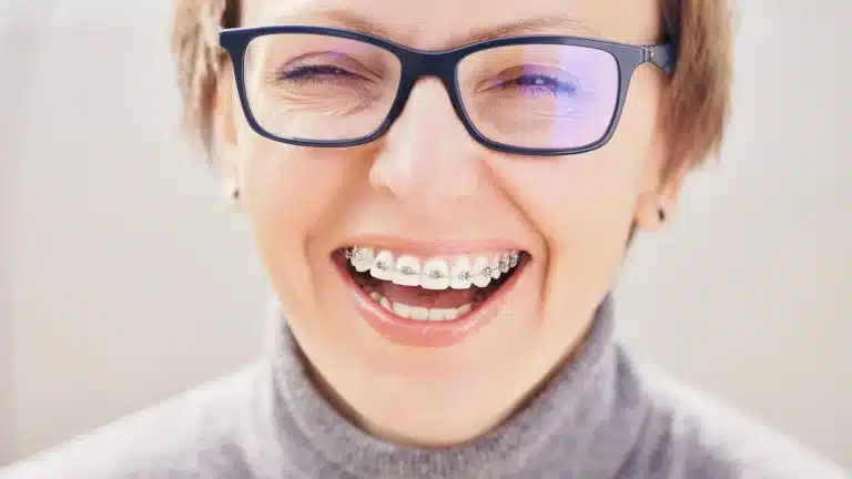 Adult wearing metal braces and smiling while looking at the camera with black-framed glasses.